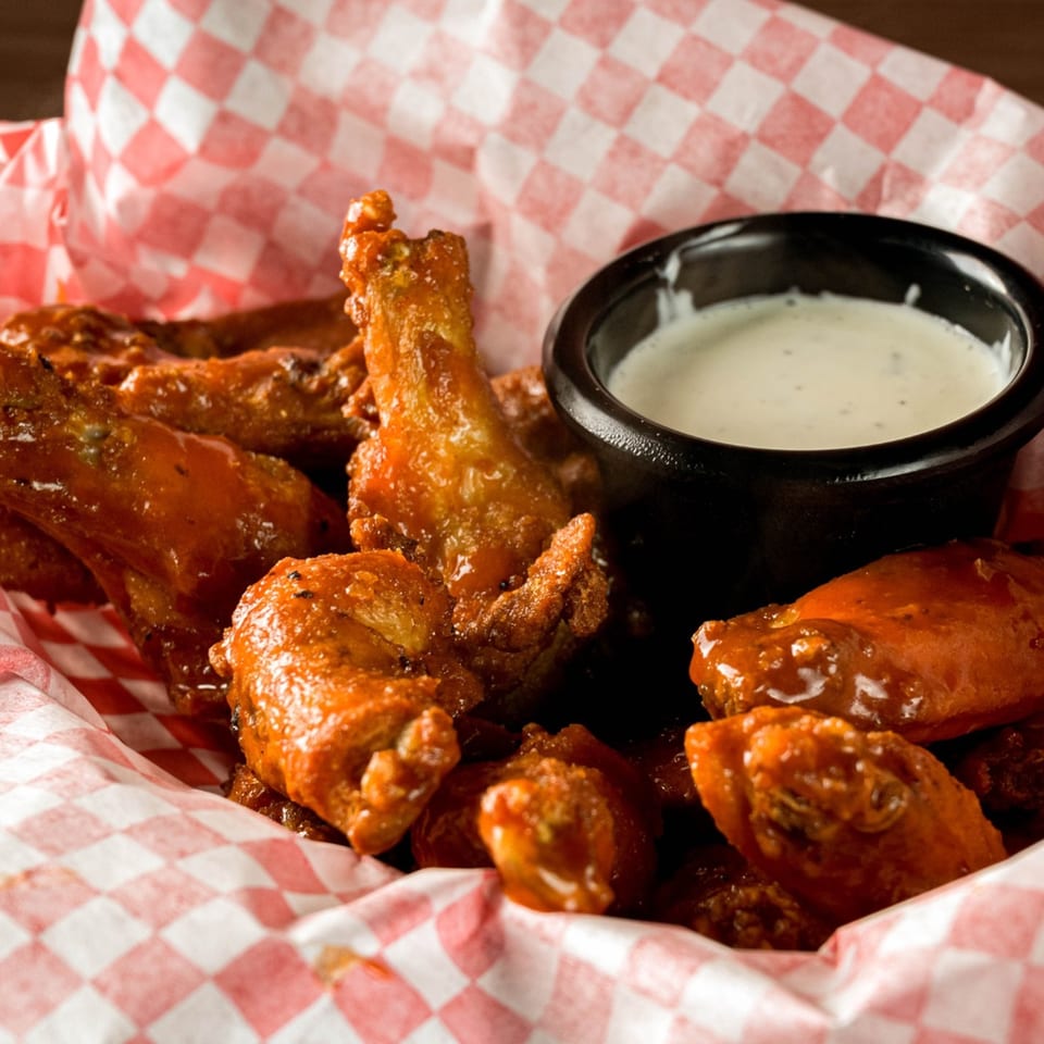 Basket of saucy chicken wings with a side of ranch dressing, served in red and white checkered paper.