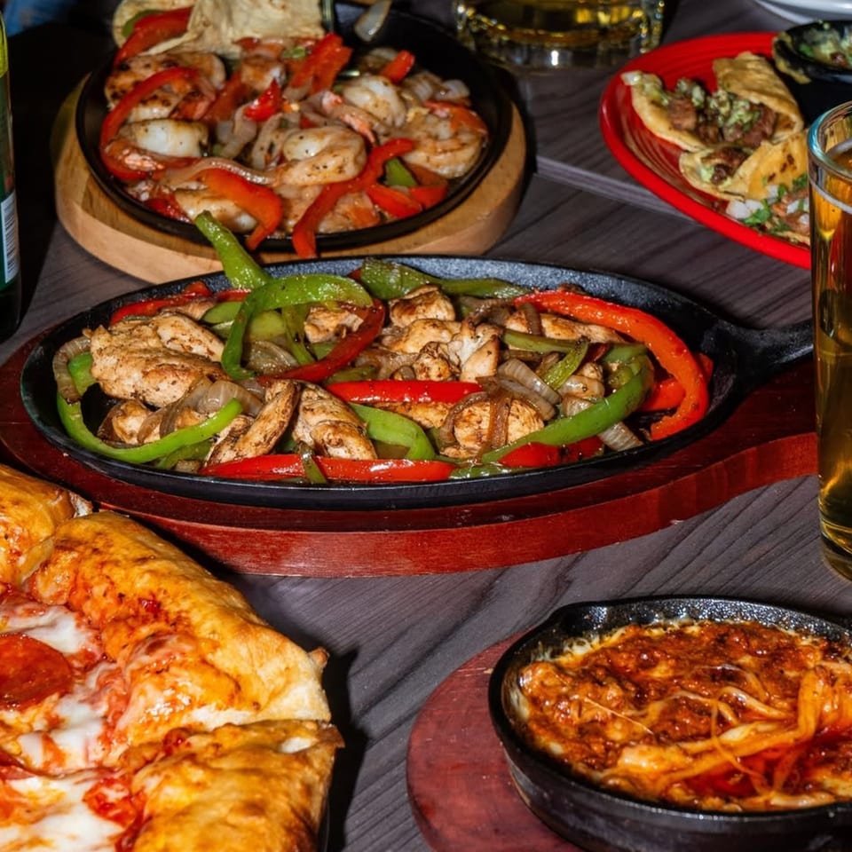 Table laden with sizzling fajitas, tacos, pepperoni pizza, and a cheesy dip, with drinks.