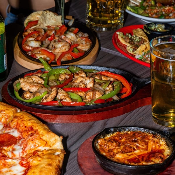 Sizzling fajitas, tacos, a pepperoni pizza, and a cheesy dip on a dark wooden table.