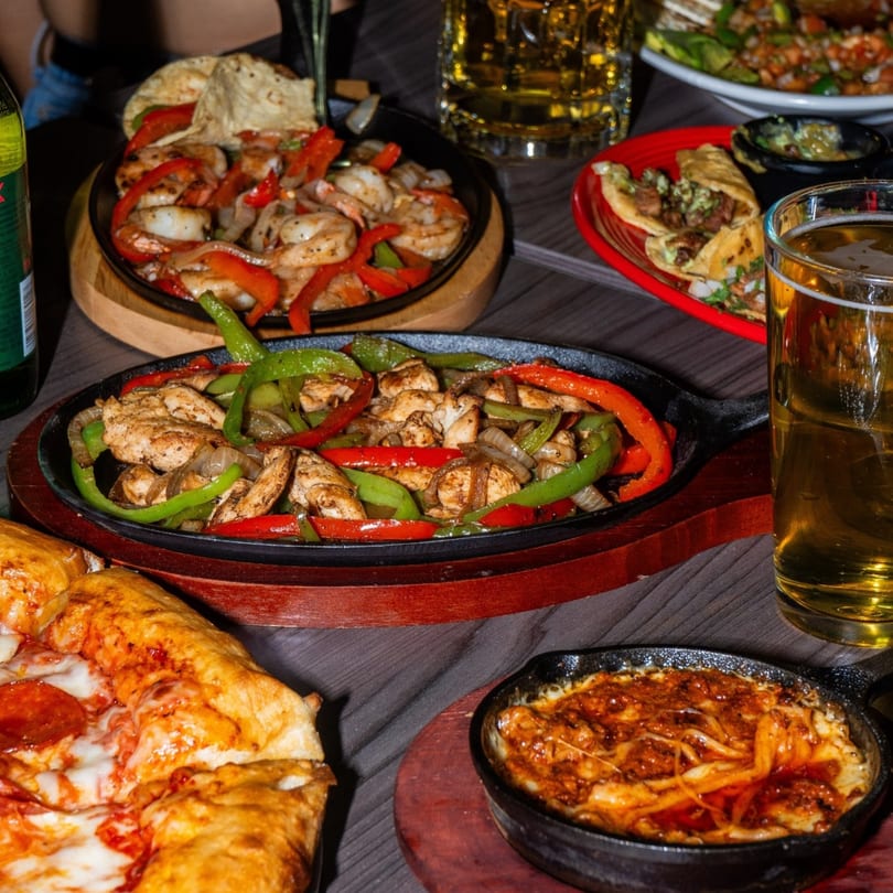 Sizzling fajitas, tacos, a pepperoni pizza, and a cheesy dip on a dark wooden table.