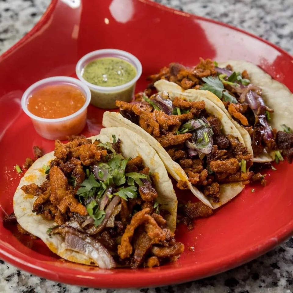Three tacos with meat, onions, and cilantro on a red plate, with two small sauce cups.