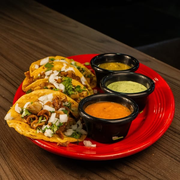 Four tacos with meat, onions, and cilantro on a red plate, with three small bowls of sauces.