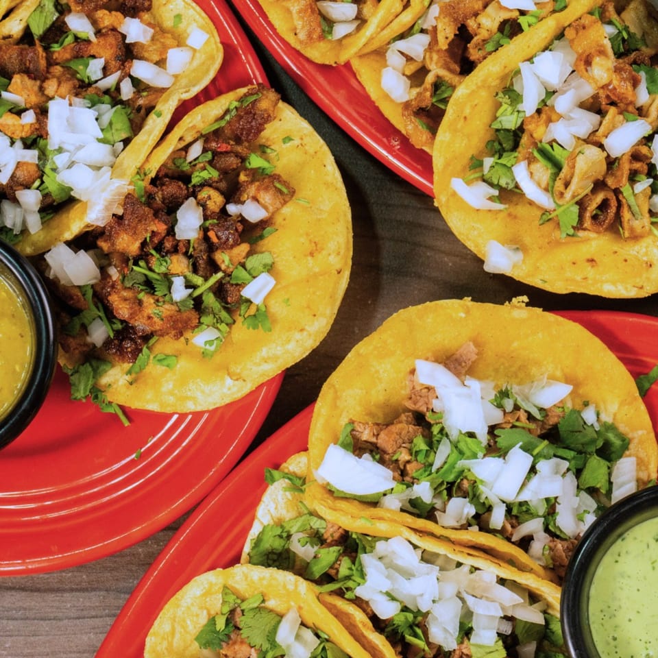 Several street tacos with meat, cilantro, and diced onions on red plates with green sauce.