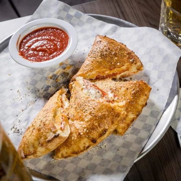 Golden-brown calzones, dusted with seasoning, served with a side of red marinara sauce on a metal tray.