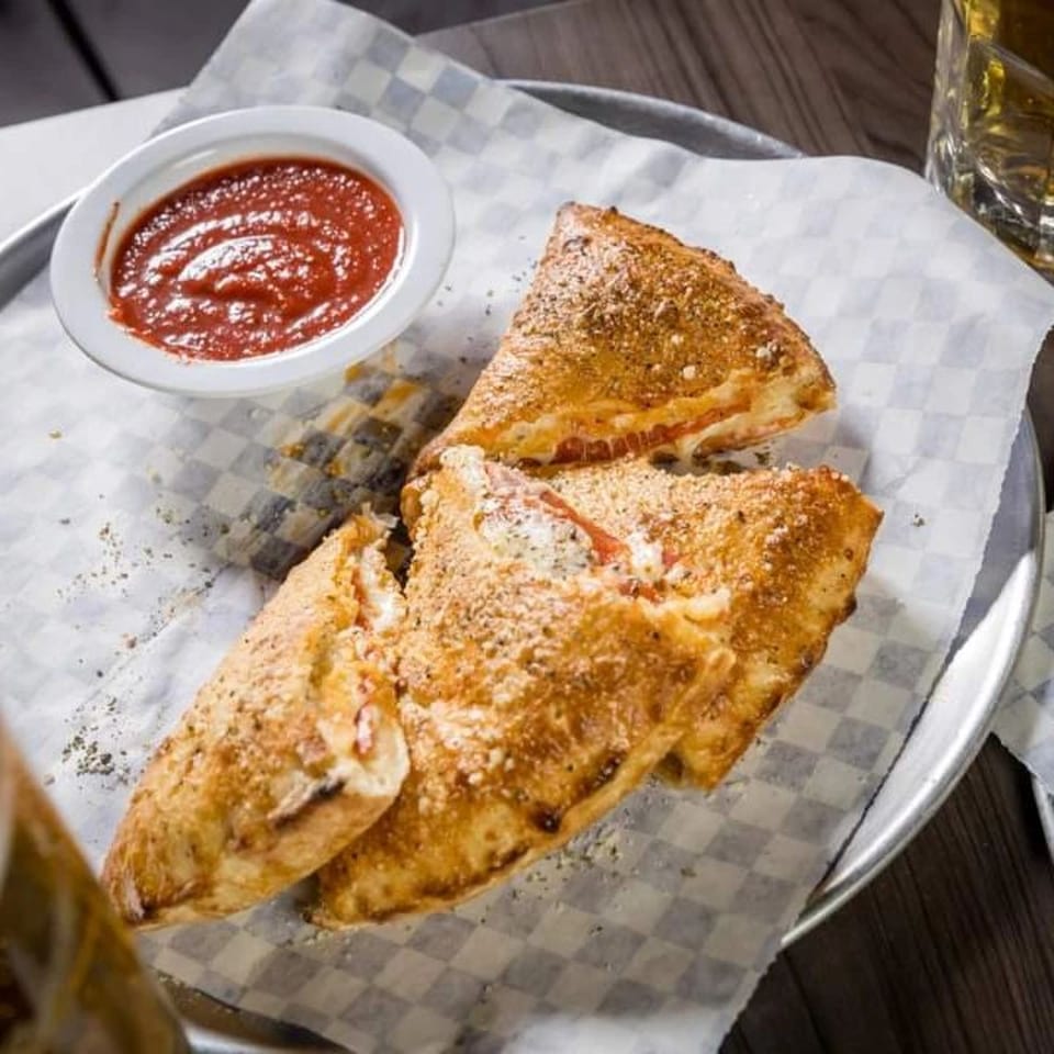 Golden-brown calzones, dusted with seasoning, served with a side of red marinara sauce on a metal tray.