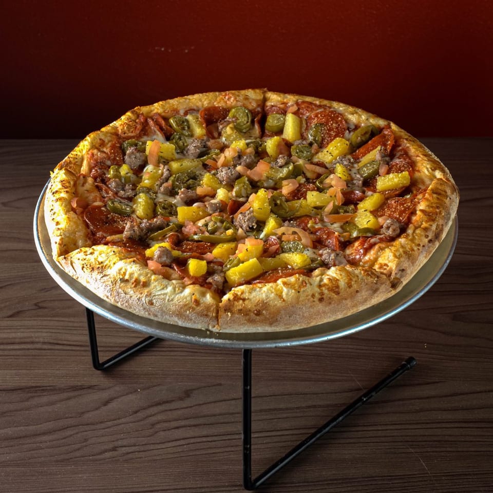 A whole Pizxa Zone pizza with pepperoni, sausage, pineapple, and jalapeños on a metal stand.