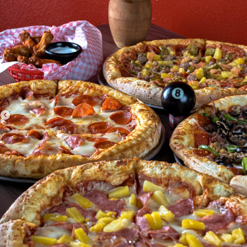 Four pizzas with various toppings, including pepperoni, pineapple, and sausage, alongside chicken wings.