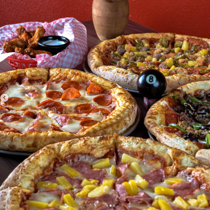 Four pizzas with various toppings, including pepperoni, pineapple, and sausage, alongside chicken wings.